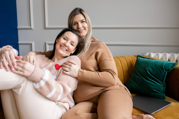 Happy women in textile suits sit on stylish mustard color sofa hugging each other in living room. Friends have fun and relax on female meeting. Home leisure, lifestyle, only women. - Foto, Imagem