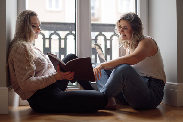 Boldog gyönyörű nők ülnek együtt a padlón, közel az erkélyhez, beszélgetni és könyvet olvasni. Barátok portréja, amint nevetnek és pihennek. Otthoni szabadidő, életmód, csak nők, női beszélgetés. - Fotó, kép