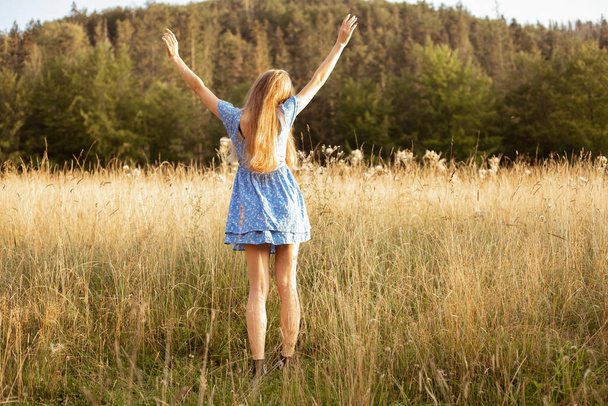 Вид сзади рыжеволосой расслабленной женщины в синем летнем платье, стоящей с поднятыми руками в солнечном поле с цветами на фоне зеленого леса в сельской местности. Сельский уютная среда и свобода в природе - Фото, изображение