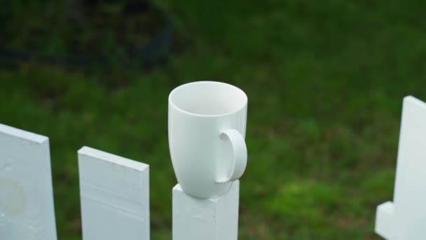 Uma xícara de cerâmica branca fica em uma cerca de madeira branca contra um fundo borrado. Café da manhã fora da cidade, paralaxe. Imagens FullHD de alta qualidade - Filmagem, Vídeo