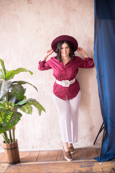 A young girl stylist with dark hair smiles in a trendy burgundy color shirt and light trousers and a wide-brimmed burgundy hat in a bright studio with green living plants in pots - Photo, Image