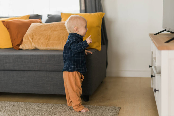 Малыш смеется, стоя рядом с диваном в гостиной. Очаровательный ребенок делает первые шаги в одиночку. Счастливого детства и ухода за детьми - Фото, изображение