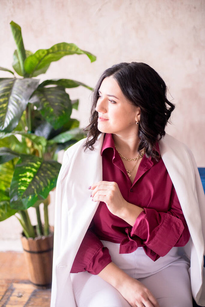 Eine junge Stylistin mit dunklen Haaren lächelt in einer trendigen weißen Jacke, einem weinroten Hemd und einer hellen Hose in einem hellen Studio mit grünen lebenden Pflanzen in Töpfen. - Foto, Bild
