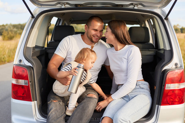 Outdoor shot of cheerful family traveling together by car, drinking tea or coffee from thermos while sitting in automobile trunk, expressing love and happiness. - Foto, afbeelding