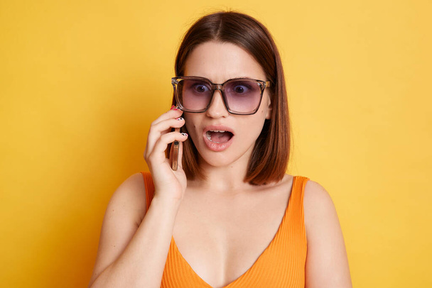 Portrait of shocked brown haired young woman wearing summer clothing talking on smart phone, hearing bad news, keeps mouth open, standing isolated over yellow background - Фото, зображення