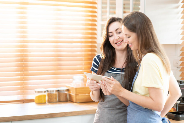 Beautiful lesbian couple cooking healthy food, looking on digital tablet at modern kitchen interior. Homosexual relations and healthy eating concept.  LGBTQ+,LGBT,LGBTQ home online kitchen. - Photo, Image
