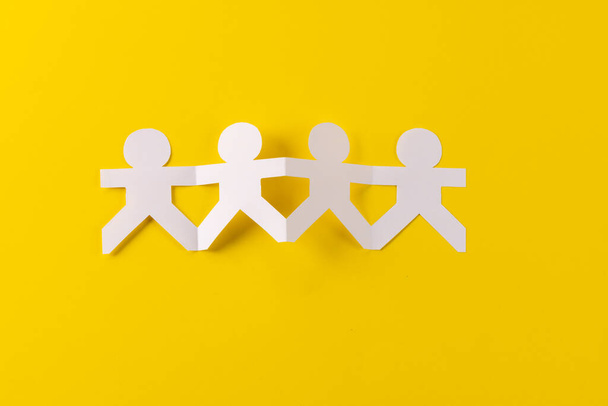 Close up of four paper cut out people figures holding hands with copy space on yellow background. Humanitarian, people, help and human concept. - Photo, Image