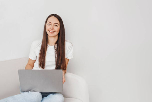 jong Kaukasisch brunette vrouw glimlachen op camera en zitten met laptop op bank kopiëren ruimte voor tekst - Foto, afbeelding