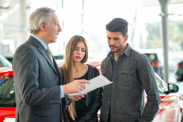 Young family talking to the salesman and choosing their new car in a showroom - Foto, Imagen