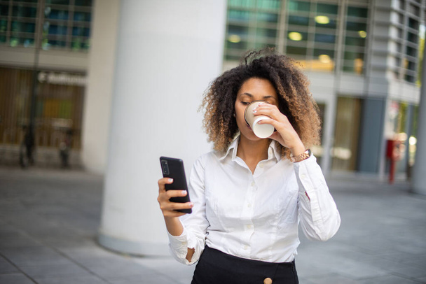 Portret afrykańskiej biznesmenki korzystającej z telefonu komórkowego w środowisku biznesowym podczas picia kawy - Zdjęcie, obraz