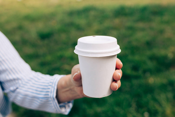 Womans hand holding white paper cup of coffee on natural morning background. Place for logo. Coffee to go. Disposable paper cup closeup. Blank space for text, mockup - Фото, зображення