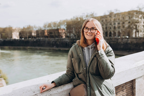 Beautiful smiling female talking on Phone and standing outdoors. Phone Communication. Happy cheerful young woman walking on city street, Urban lifestyle concept. - 写真・画像