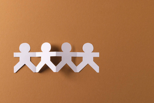 Close up of four paper cut out people figures holding hands with copy space on brown background. Humanitarian, people, help and human concept. - Photo, Image