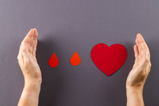 Hands of caucasian woman protecting blood drops and heart, on grey background. Blood donation, heart health, medicine and healthcare. - Foto, imagen