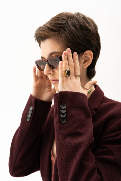 brunette woman in burgundy brown blazer and finger rings adjusting stylish sunglasses isolated on white - Foto, Imagen