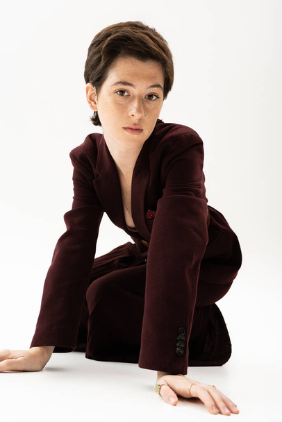 young model with short brunette hair posing in brown pantsuit and looking at camera on white background - Foto, Bild