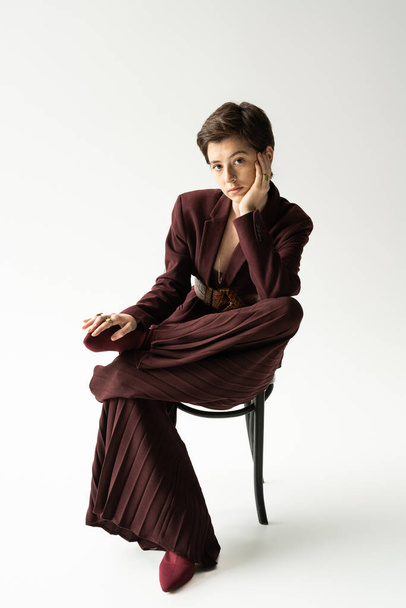 young and elegant woman in wide pants looking at camera while posing on chair on grey background - Valokuva, kuva