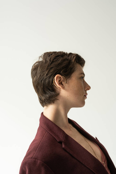profile of stylish brunette woman in cuff earring and blazer isolated on grey - Foto, Bild