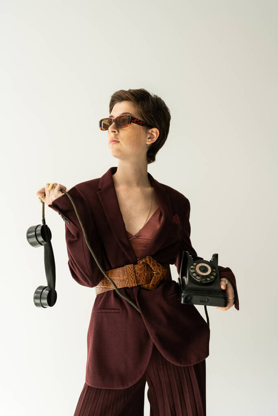 mulher morena em óculos de sol e casaco marrom borgonha com cinto de couro posando com telefone vintage isolado em cinza - Foto, Imagem