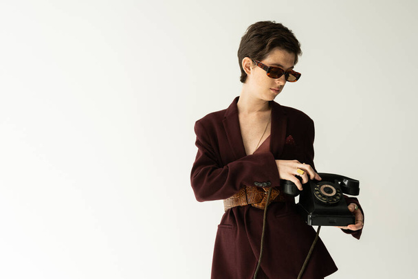 брюнетка в модном пиджаке с кожаным ремнем, стоящим с винтажным телефоном на сером фоне - Фото, изображение