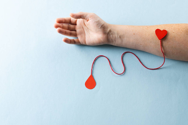 Arm of caucasian woman with heart and red string with blood drop, on blue background. Blood donation, medicine and healthcare. - Фото, изображение