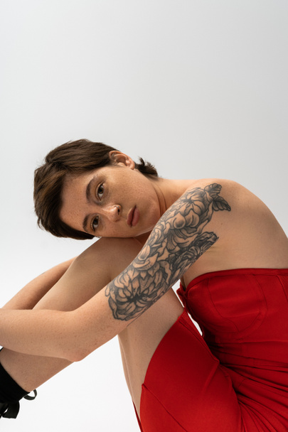 mujer tatuada de ensueño en vestido de corsé rojo sentado y mirando a la cámara sobre fondo gris - Foto, Imagen
