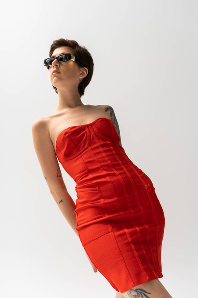 low angle view of slender woman in red strapless dress and stylish sunglasses on grey background - Foto, afbeelding