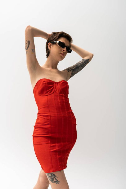 vista de ángulo bajo de mujer tatuada en gafas de sol y vestido de corsé rojo posando con las manos detrás de la cabeza en gris  - Foto, Imagen