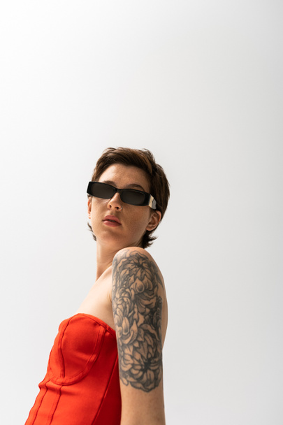brunette tattooed woman in sunglasses and red corset dress looking at camera on grey background - Φωτογραφία, εικόνα