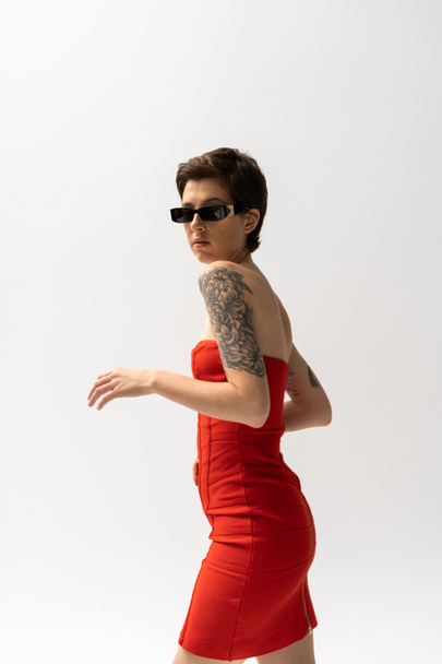 barna tetovált nő napszemüveg és piros fűző ruha nézi kamera elszigetelt szürke - Fotó, kép