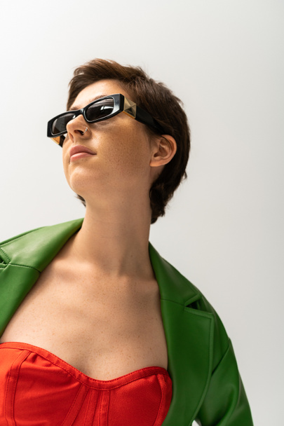 low angle view of young brunette woman in sunglasses and green leather jacket looking away isolated on grey - Photo, Image