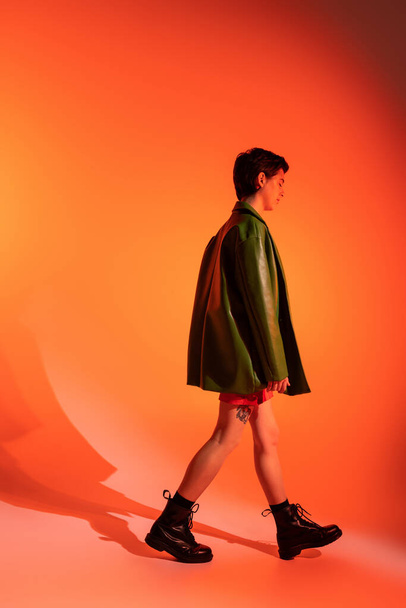 side view of tattooed woman in green jacket and black boots walking on orange background - Foto, Imagen