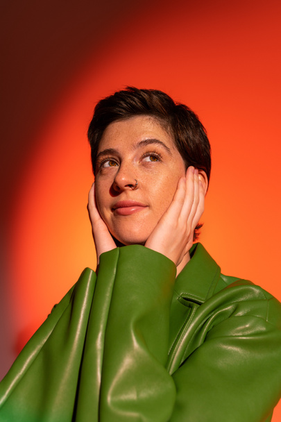 dreamy and positive woman in green leather jacket touching face and looking away on orange shaded background - 写真・画像