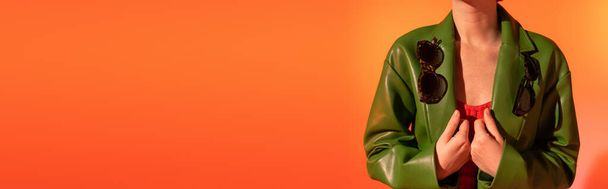 Yeşil deri ceketli, son moda güneş gözlüklü, turuncu arka planda poz veren kadın manzarası, afiş - Fotoğraf, Görsel