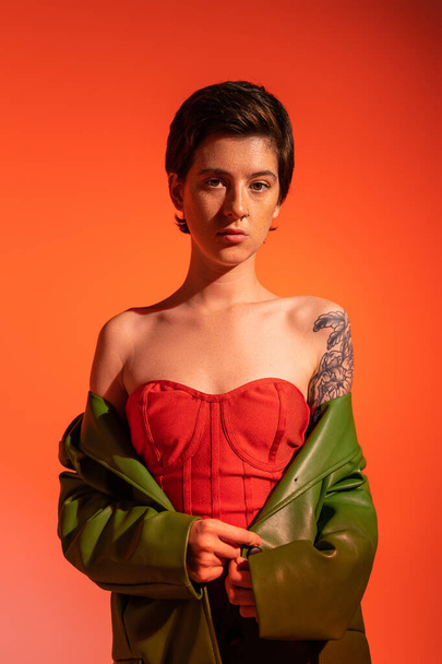 young tattooed woman in red corset dress and green leather jacket looking at camera on orange background - Foto, imagen