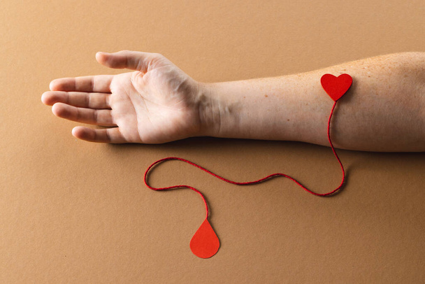 Arm of caucasian woman with heart and red string with blood drop, on brown background. Blood donation, medicine and healthcare. - Photo, Image