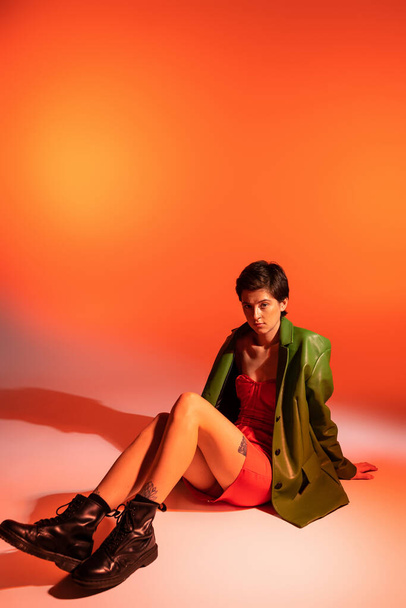 full length of fashionable brunette woman in green jacket and black leather boots sitting on orange background - Fotó, kép