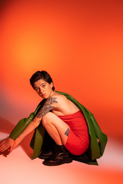 full length of tattooed woman in red corset dress and black boots sitting on haunches and looking at camera on orange and pink background - 写真・画像