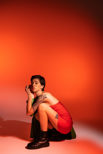 mujer tatuada de moda en vestido rojo sin tirantes y botas de cuero negro posando sobre melocotones sobre fondo naranja - Foto, Imagen