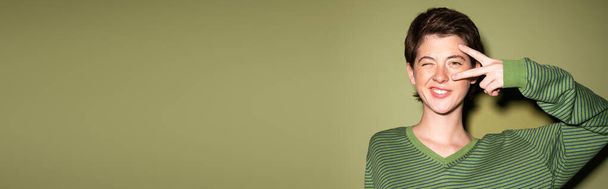 donna bruna sorridente strizzando l'occhio alla macchina fotografica e mostrando il segno della pace vicino al viso su sfondo verde, banner - Foto, immagini