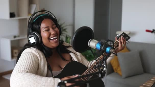Young woman sitting at table with microphone and singing song while playing guitar - curvy influencer girl, recording herself streaming for her audience - podcast, social media - - Footage, Video