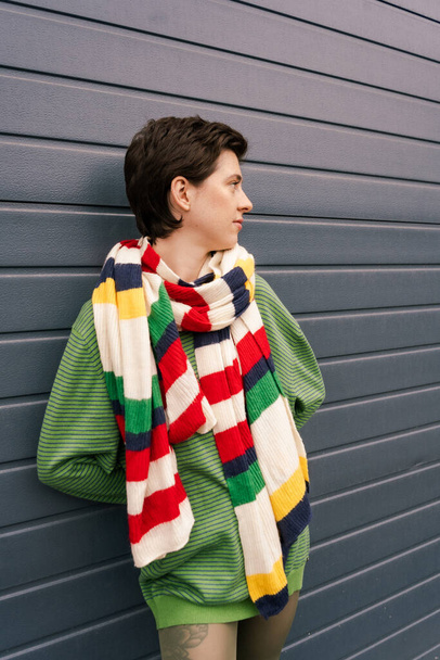 brunette woman in striped scarf looking away near grey wall on city street - Foto, imagen