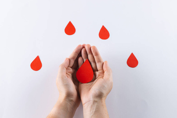 Hands of caucasian woman cupping blood drop, with blood drops on white background. Blood donation, medicine and healthcare. - Φωτογραφία, εικόνα