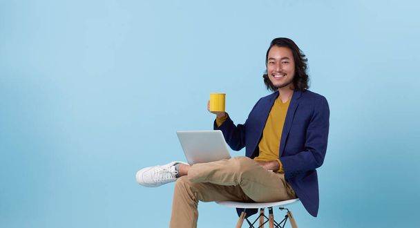 Jovem asiático homem de negócios feliz sorrindo usando computador portátil sentado na cadeira branca e beber café isolado no fundo do estúdio azul. - Foto, Imagem