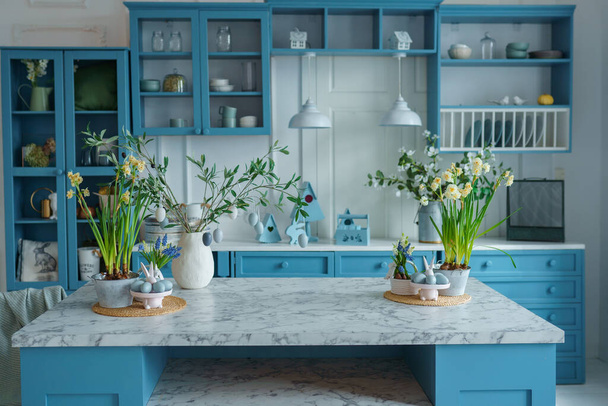 Blaue Kücheneinrichtung mit Möbeln. Stilvolle Küche mit Blumen in der Vase. Holzküche im Frühlingsdekor. Gemütliche Wohnkultur. Küchenutensilien, Geschirr und Teller auf dem Tisch. Kücheninsel im Esszimmer, Hasenfigur. Osterkonzept - Foto, Bild
