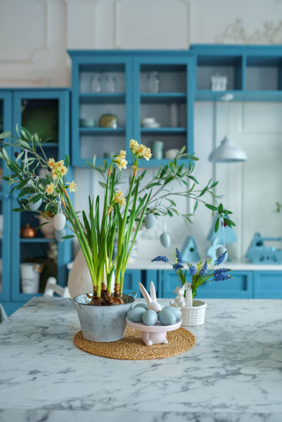 Niebieskie wnętrze kuchni z meblami. Stylowa kuchnia z kwiatami w wazonie. Drewniana kuchnia w wiosennym wystroju. Przytulny wystrój domu. Przybory kuchenne, naczynia i talerz na stole. wyspa kuchnia w jadalni, figurka króliczka. Koncepcja wielkanocna - Zdjęcie, obraz