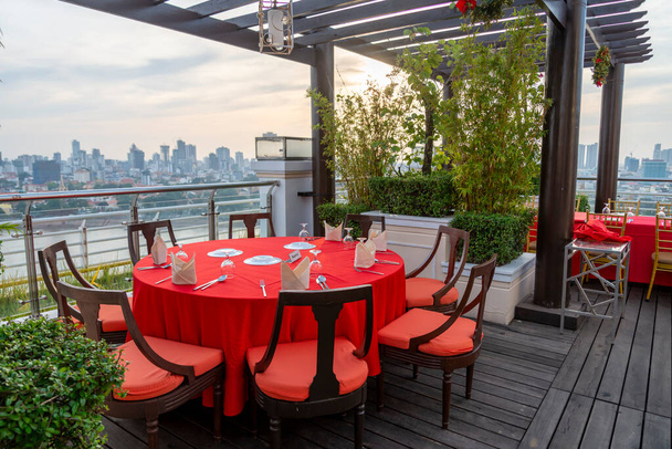 Leuchtend rote Tischdecke, die über einer runden Holztischplatte hängt, mit silbernem Besteck, Tellern und Servietten bestückt und von acht Luxusstühlen mit roten Kissen umgeben. - Foto, Bild