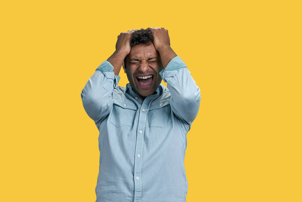 Young indian man yelling with aggressive expression and hands holding head. Lose and frustration concept. Isolated on yellow. - Foto, immagini