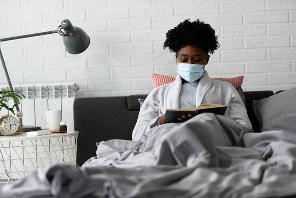 Αφροαμερικανή άρρωστη γυναίκα με μάσκα που κάθεται στο κρεβάτι και διαβάζει βιβλίο. - Φωτογραφία, εικόνα