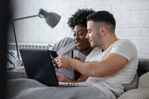 smiling mixed race couple lying on bed with laptop in bedroom at home  - Fotografie, Obrázek